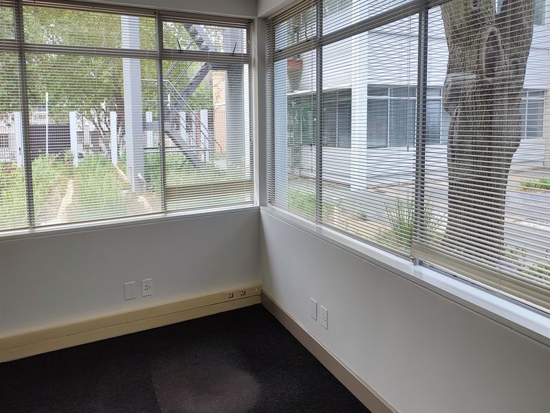 A bright, empty office space featuring large windows with venetian blinds, carpeted flooring, and integrated electrical trunking along the baseboards.