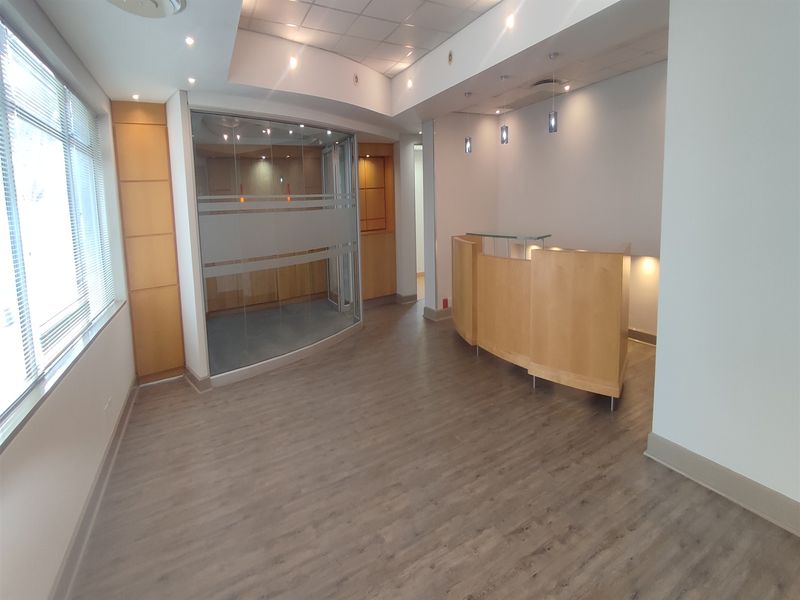 A professional office space featuring a built-in reception desk, glass partition, wood veneer accents, and modern laminate flooring with recessed lighting.