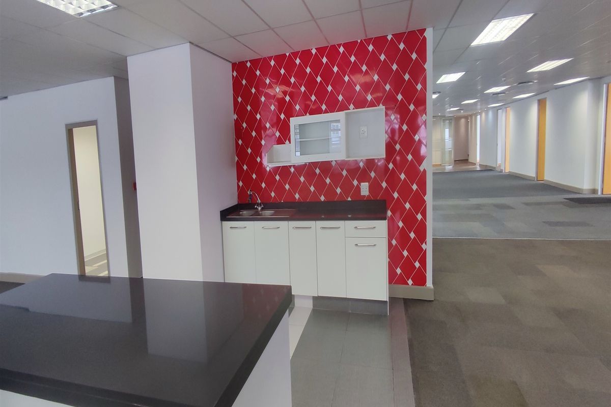 A compact office kitchenette featuring white cabinetry, a stainless steel sink, and a distinctive red patterned splashback within an open-plan office environment.
