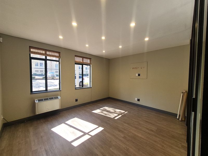 A bright, empty office space featuring modern laminate flooring, recessed ceiling lighting, and a wall-mounted HVAC unit with large windows.
