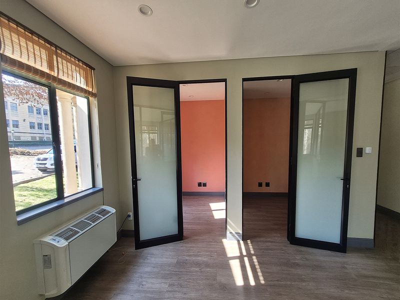 A bright, empty office space featuring modern laminate flooring, recessed lighting, an integrated HVAC unit, and double frosted glass doors leading to adjacent rooms.