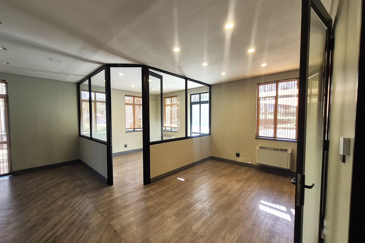 A bright, modern office space featuring laminate flooring, recessed lighting, and a glass-partitioned private office area with an integrated HVAC unit.