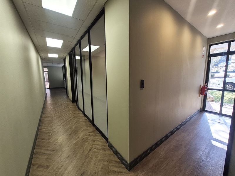 Modern office hallway featuring herringbone vinyl flooring, glass partition walls, and recessed lighting, leading to an exterior entrance with natural light.