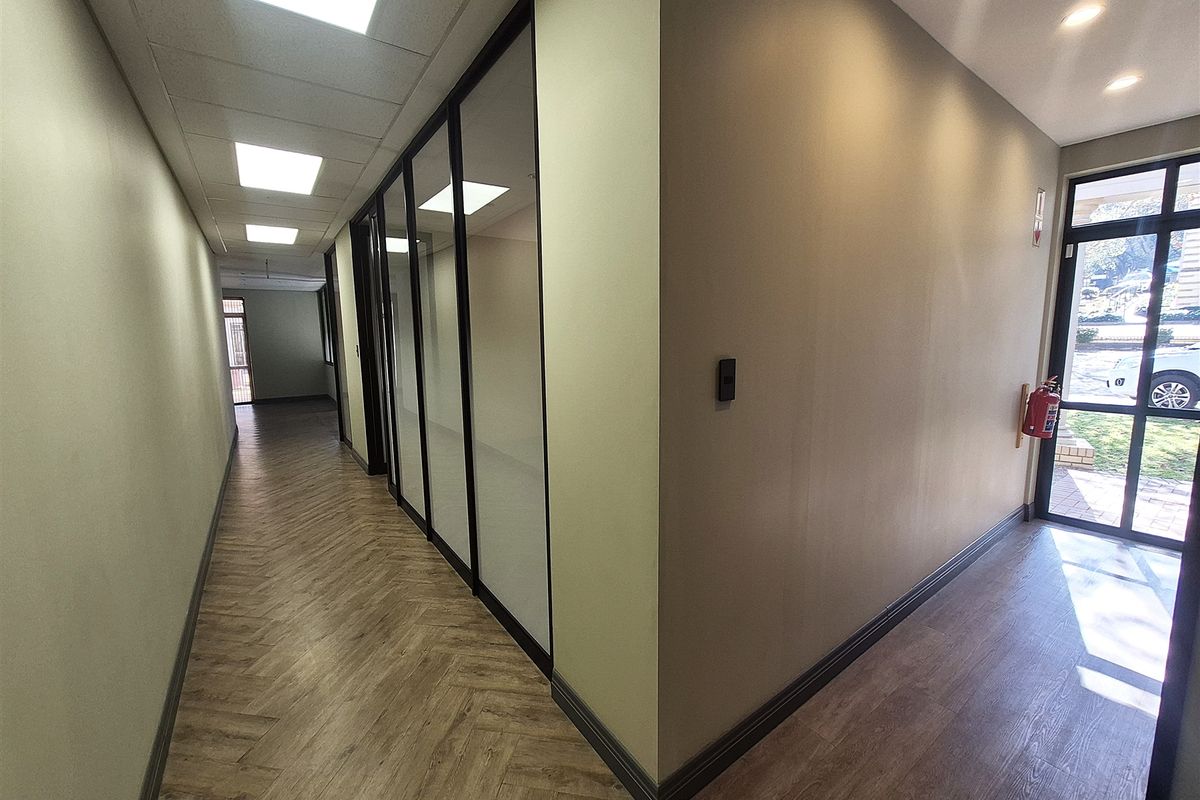Modern office hallway featuring herringbone vinyl flooring, glass partition walls, and recessed lighting, leading to an exterior entrance with natural light.