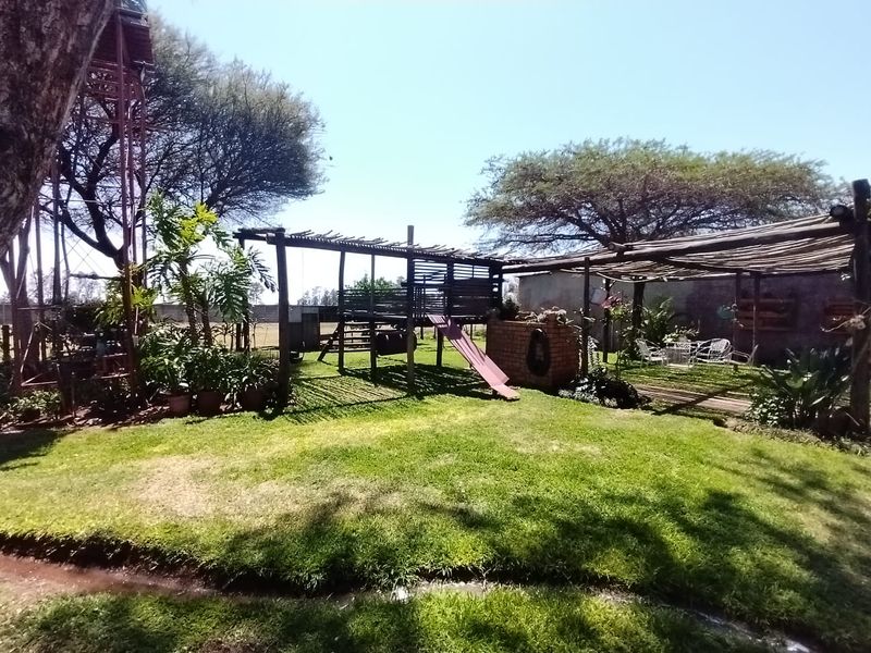 Spacious farm garden featuring a lawn, timber pergola, brick braai area, and a children's play structure with an adjacent water tower.