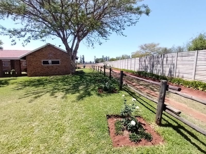 A well-maintained garden area featuring a brick farmhouse exterior, mature trees, and timber post and rail fencing along a concrete boundary wall.