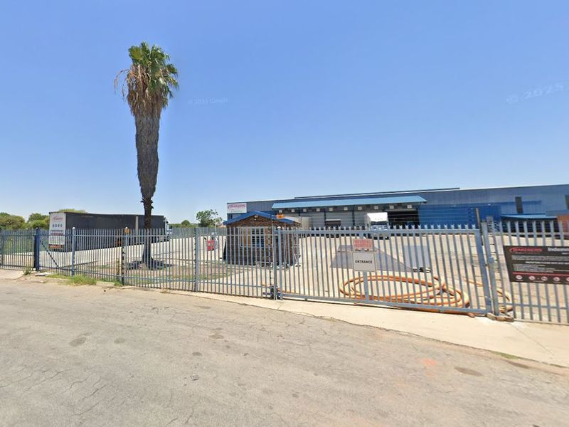 Front view of an industrial warehouse property featuring a secure gated entrance, perimeter palisade fencing, and a dedicated security guard hut.