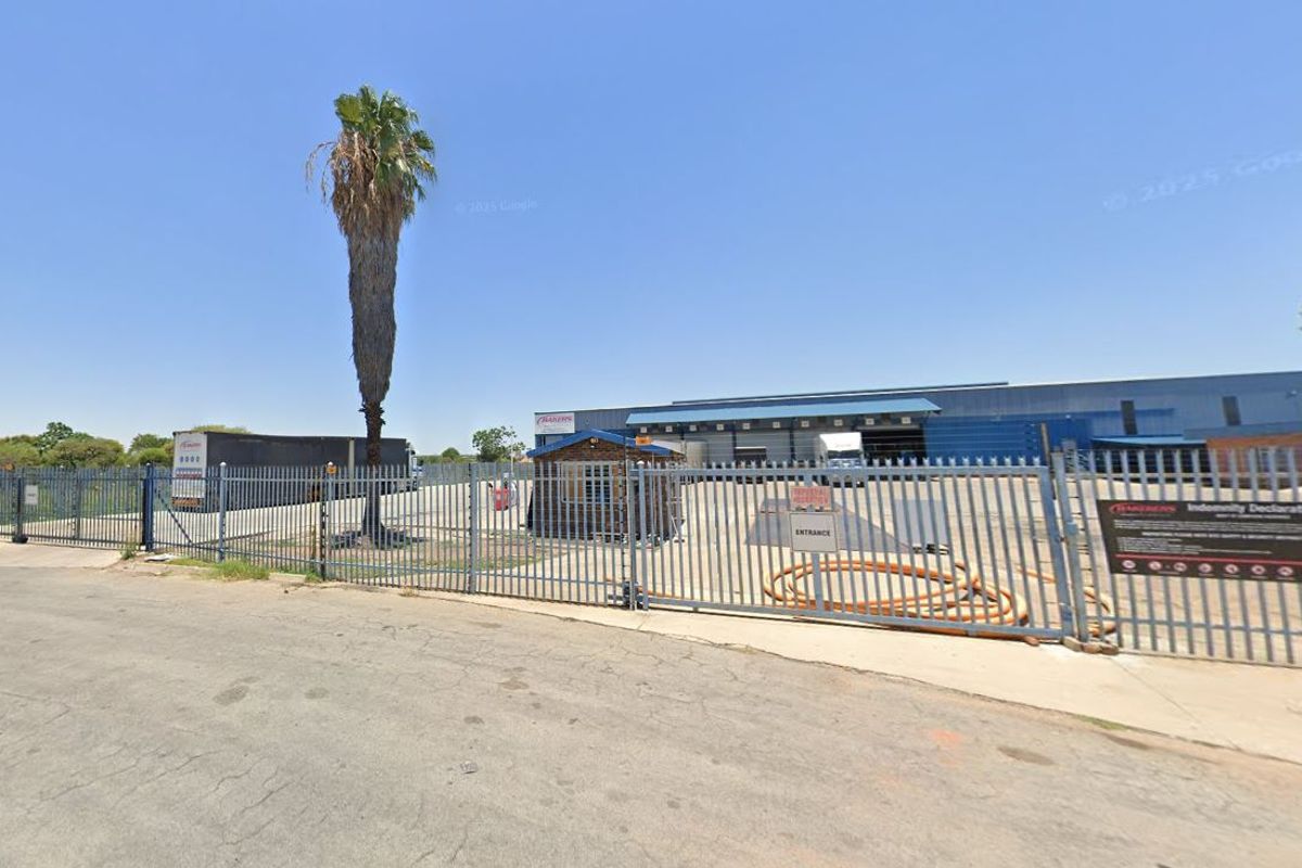Front view of an industrial warehouse property featuring a secure gated entrance, perimeter palisade fencing, and a dedicated security guard hut.