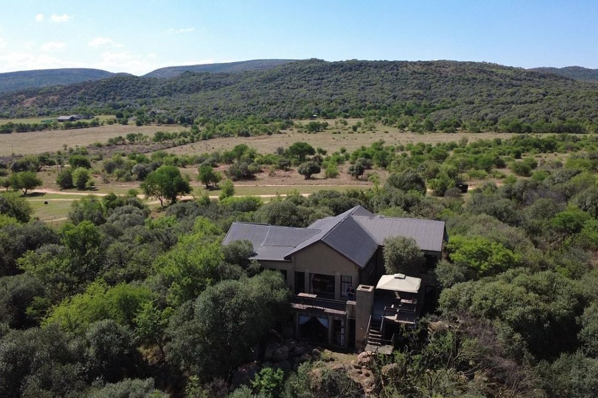 Aerial view of a residential small holding property nestled in a wooded landscape with a metal roof and elevated outdoor deck area.
