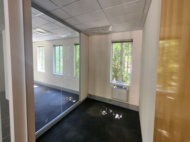 An empty office space featuring a glass partition wall, suspended ceiling, wall-mounted air conditioning unit, and carpet flooring.