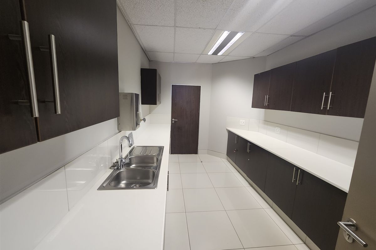 A clean, modern office kitchenette featuring dark wood cabinetry, white countertops, a double basin sink, and bright tiled flooring.