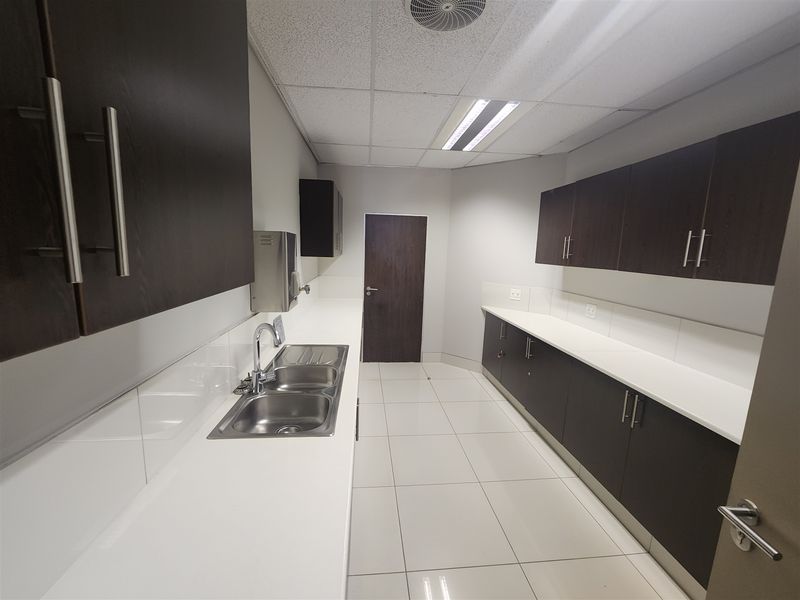 A clean, modern office kitchenette featuring dark wood cabinetry, white countertops, a double basin sink, and bright tiled flooring.