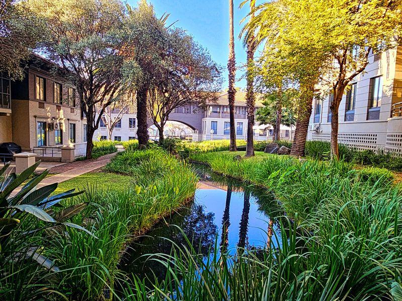 A well-maintained office complex exterior featuring a landscaped garden, a central water feature, and paved walkways between building wings.