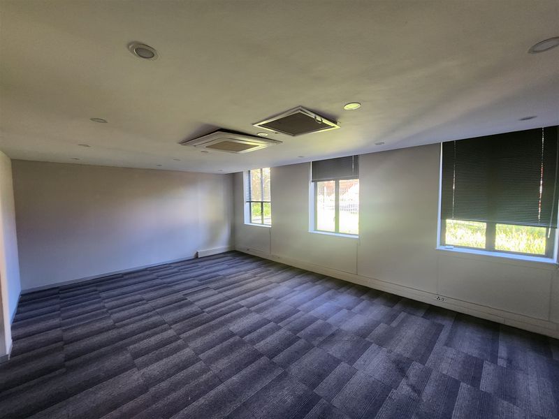 A spacious, empty office area featuring carpeted flooring, recessed lighting, ceiling-mounted air conditioning units, and large windows with fitted venetian blinds.