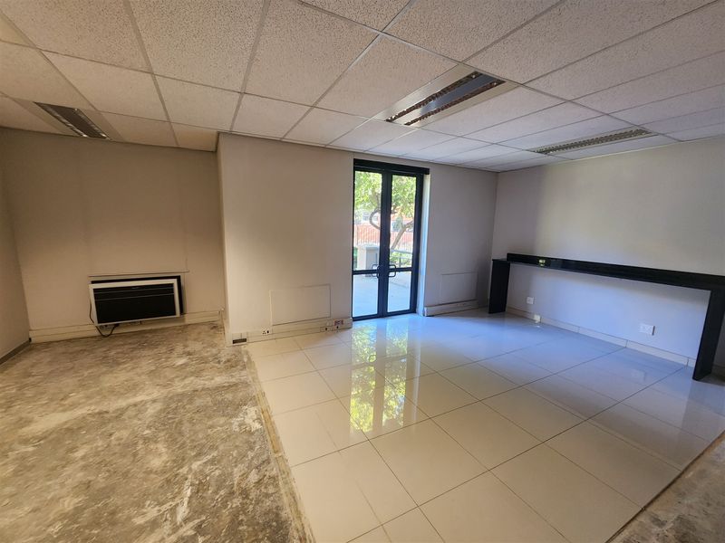 An empty office space featuring a suspended ceiling, recessed lighting, wall-mounted air conditioning, and a mix of tiled and unfinished flooring with a glass door.