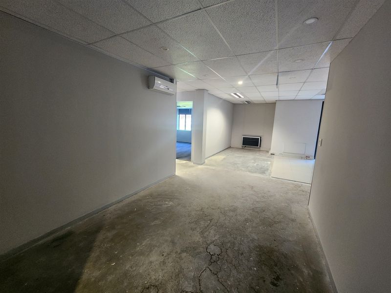 An empty office space featuring a suspended ceiling, recessed lighting, wall-mounted air conditioning, and unfinished concrete flooring ready for renovation.