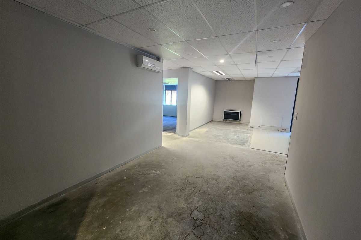 An empty office space featuring a suspended ceiling, recessed lighting, wall-mounted air conditioning, and unfinished concrete flooring ready for renovation.