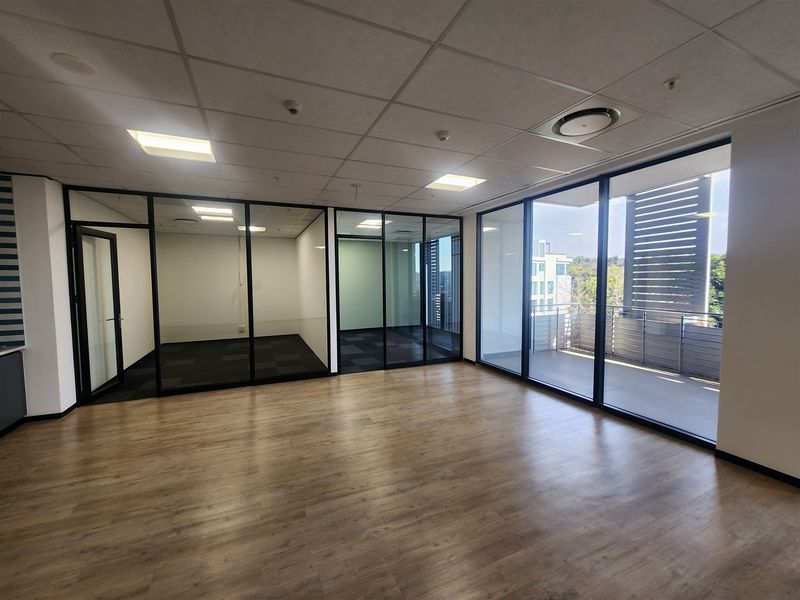 Modern open-plan office space featuring glass-partitioned meeting rooms, laminate flooring, recessed ceiling lights, and direct access to a private balcony.