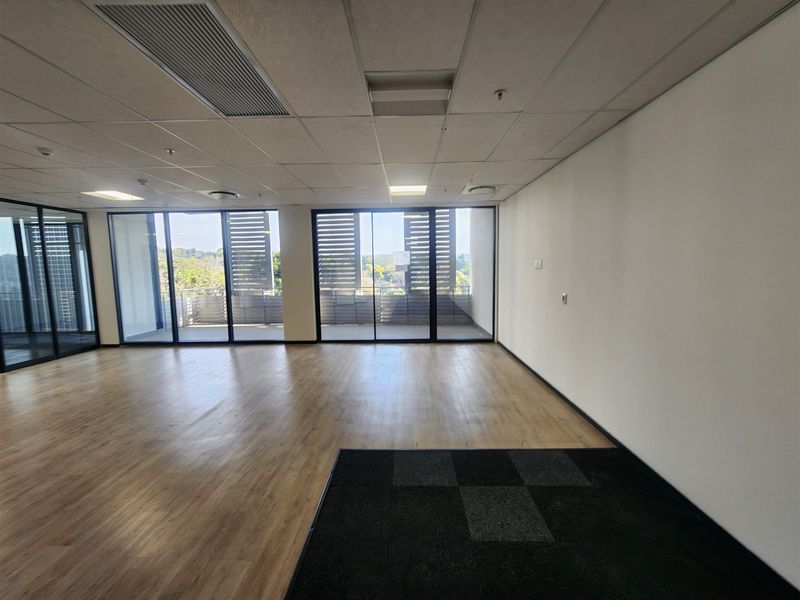 A spacious, modern open-plan office area featuring laminate flooring, carpet tile accents, suspended ceilings, and large sliding glass doors leading to an outdoor balcony.