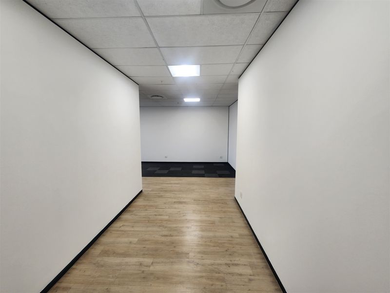 A bright, vacant office space featuring a mix of laminate and carpet flooring, suspended ceilings with recessed lighting, and clean white walls.