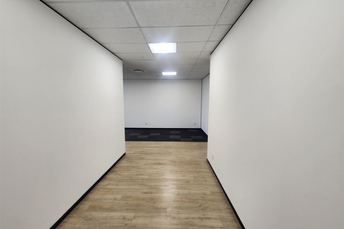 A bright, vacant office space featuring a mix of laminate and carpet flooring, suspended ceilings with recessed lighting, and clean white walls.