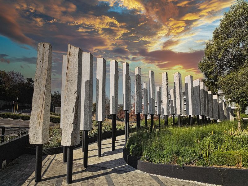 A modern landscaped exterior area featuring a row of stone pillar sculptures, paved pathways, and manicured garden beds at the office property entrance.