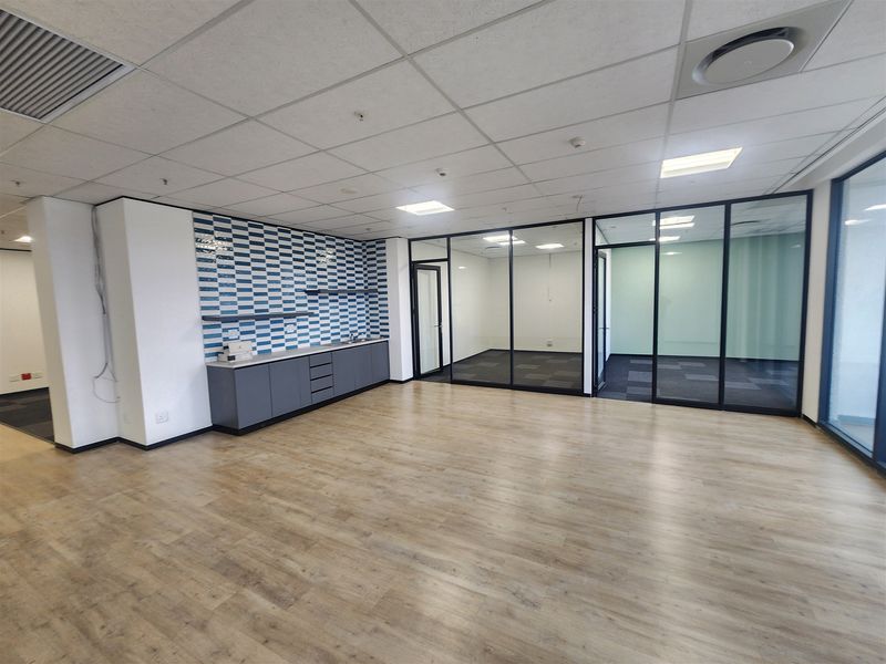 Modern open-plan office space featuring glass-partitioned meeting rooms, a built-in kitchenette, wood-look laminate flooring, and a professional suspended ceiling with recessed lighting.