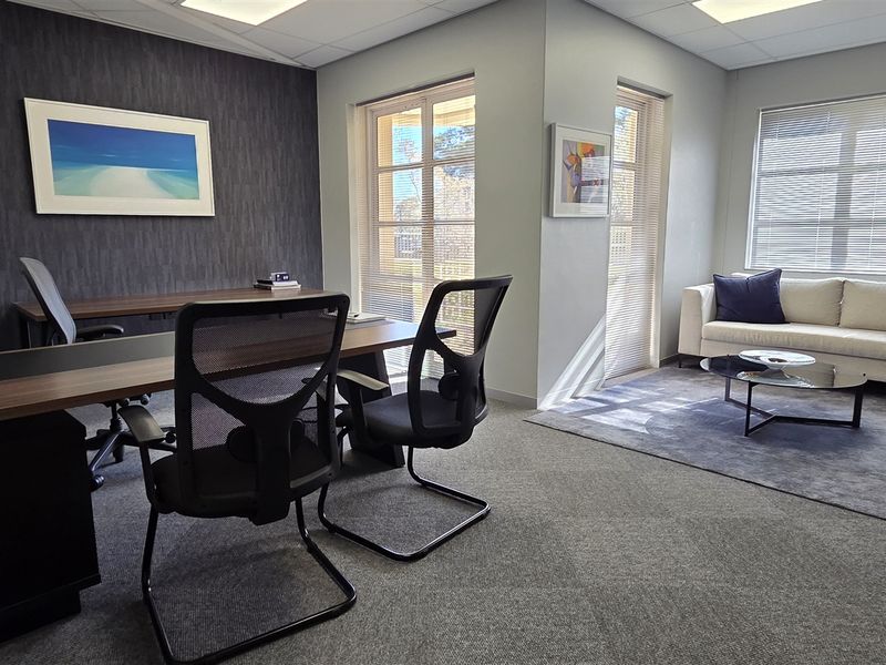 A professional office space featuring carpeted flooring, recessed ceiling lighting, and large windows with venetian blinds, creating a bright and functional work environment.