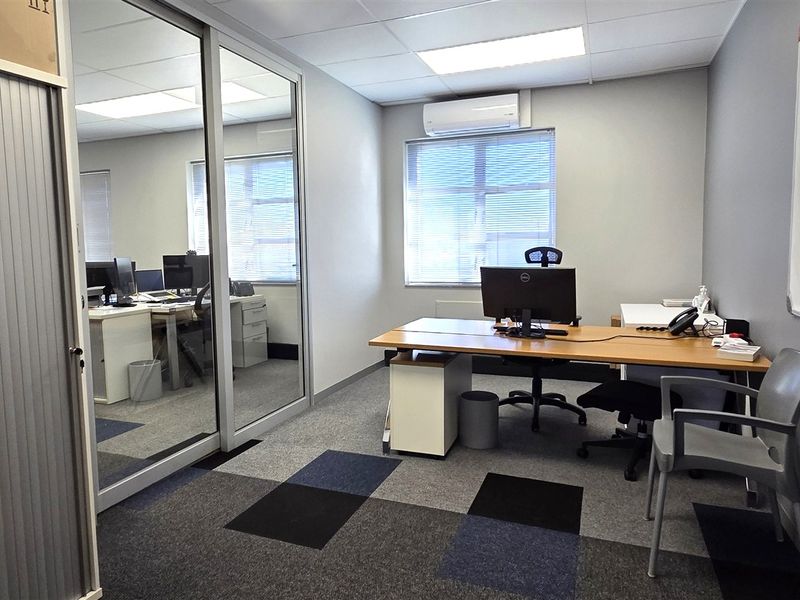 A functional office space featuring carpet tiles, a glass partition wall, a wall-mounted air conditioning unit, and a built-in whiteboard.