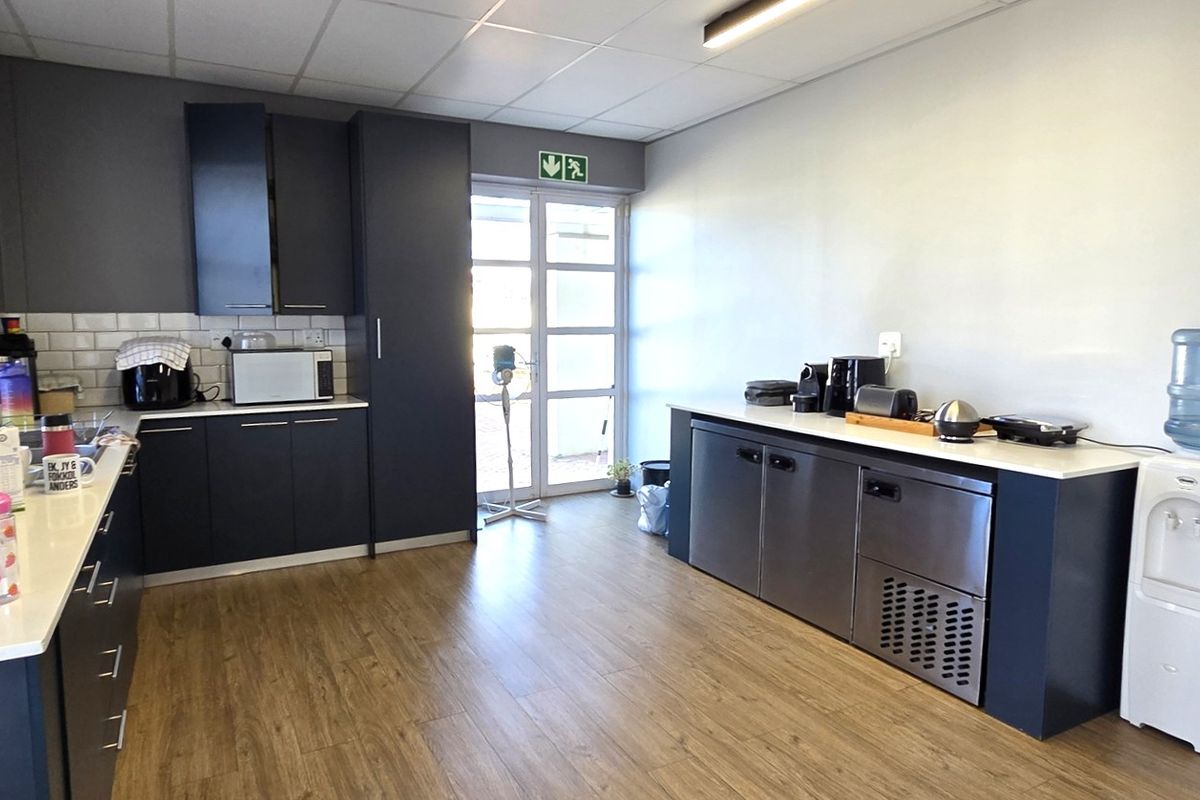 A modern office kitchen featuring dark cabinetry, white countertops, subway tile backsplash, and durable laminate flooring with direct access to an outdoor area.