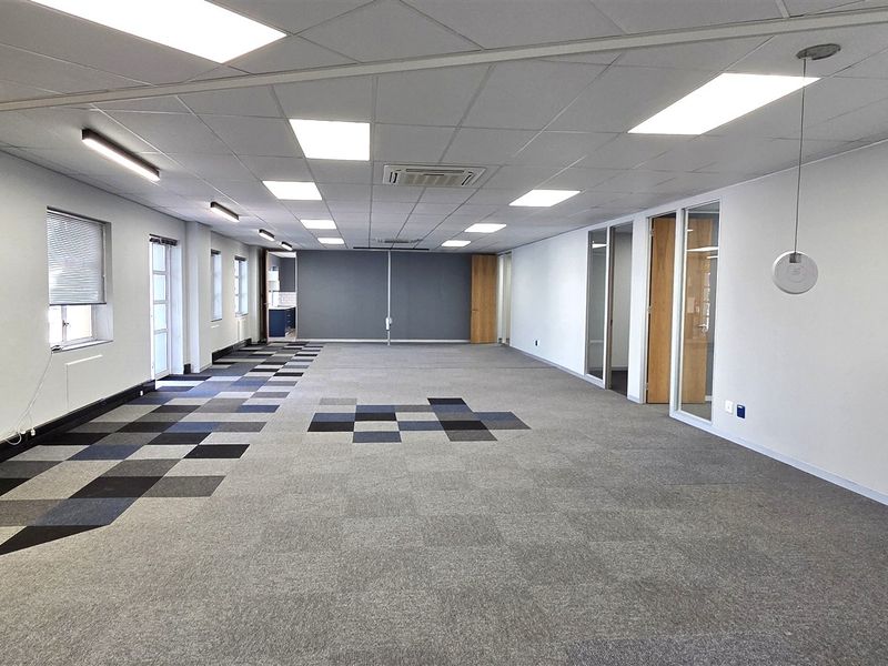A spacious, open-plan office area featuring modern carpet tiles, suspended ceilings with recessed lighting, and glass-partitioned office doors.