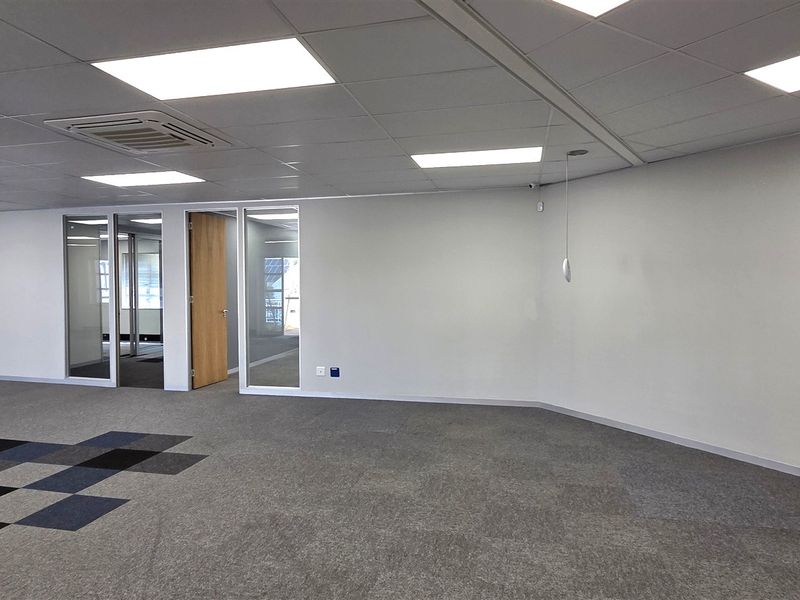 A spacious, empty open-plan office area featuring carpet tiles, suspended ceilings with recessed lighting, and glass-partitioned private office rooms.