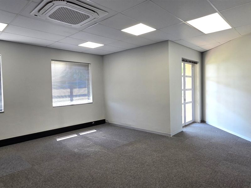 A bright, vacant office space featuring carpet tiles, a suspended ceiling with recessed LED lighting, and a ceiling-mounted air conditioning unit.