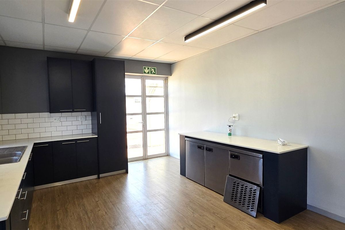 A clean, modern office kitchen area featuring dark cabinetry, white countertops, subway tile backsplash, and durable wood-look flooring with an emergency exit.