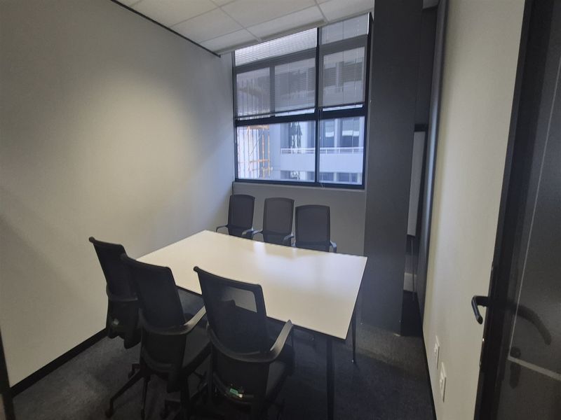 A functional office meeting room featuring carpeted flooring, a suspended ceiling, and a large window providing natural light.