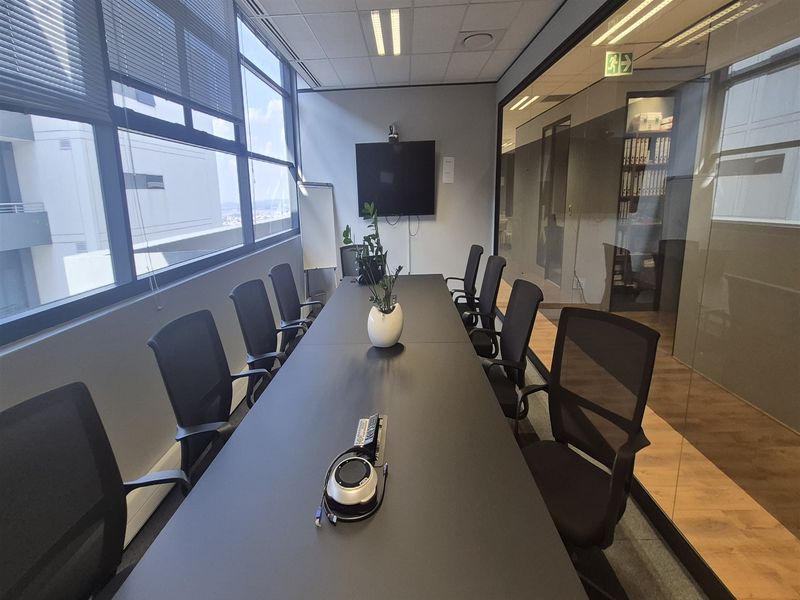 A professional office meeting room featuring a large conference table, floor-to-ceiling windows with blinds, and a modern glass partition wall.