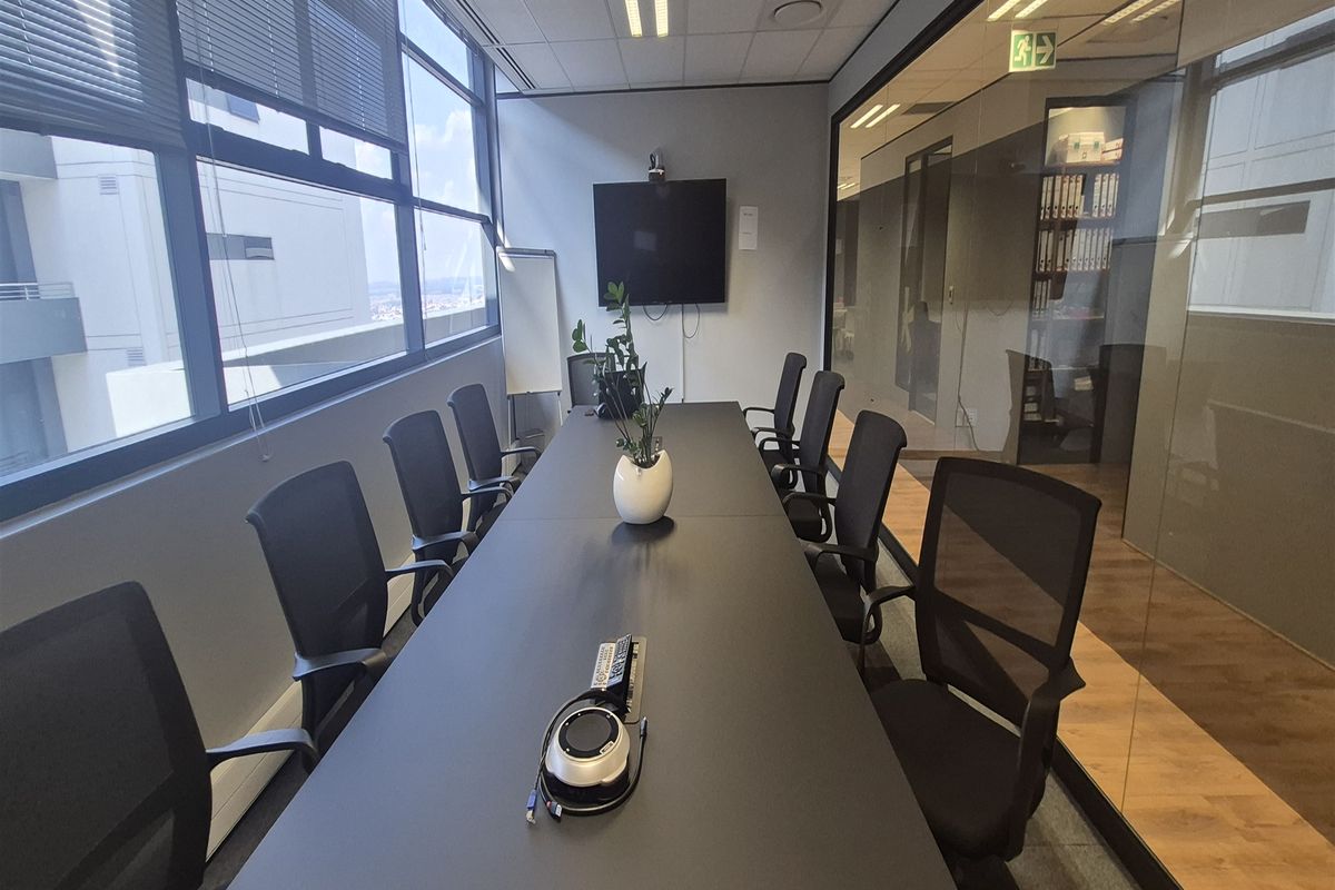 A professional office meeting room featuring a large conference table, floor-to-ceiling windows with blinds, and a modern glass partition wall.