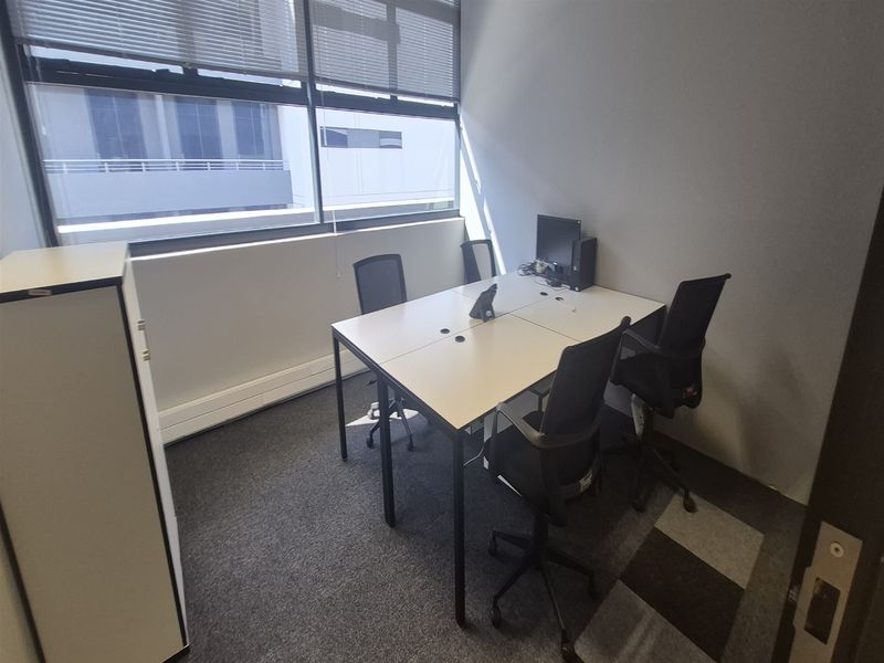 A functional office space featuring large windows with venetian blinds, carpeted flooring, and ample natural light, suitable for a professional work environment.