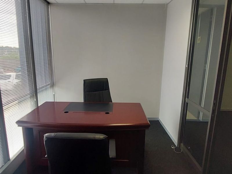A private office space featuring large windows with venetian blinds, carpeted flooring, and a glass partition door leading to an adjacent area.