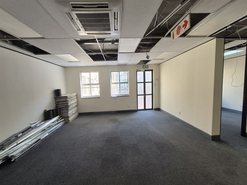 An empty office space featuring carpet flooring, a cassette air conditioning unit, and a suspended ceiling with several missing tiles and exposed wiring.