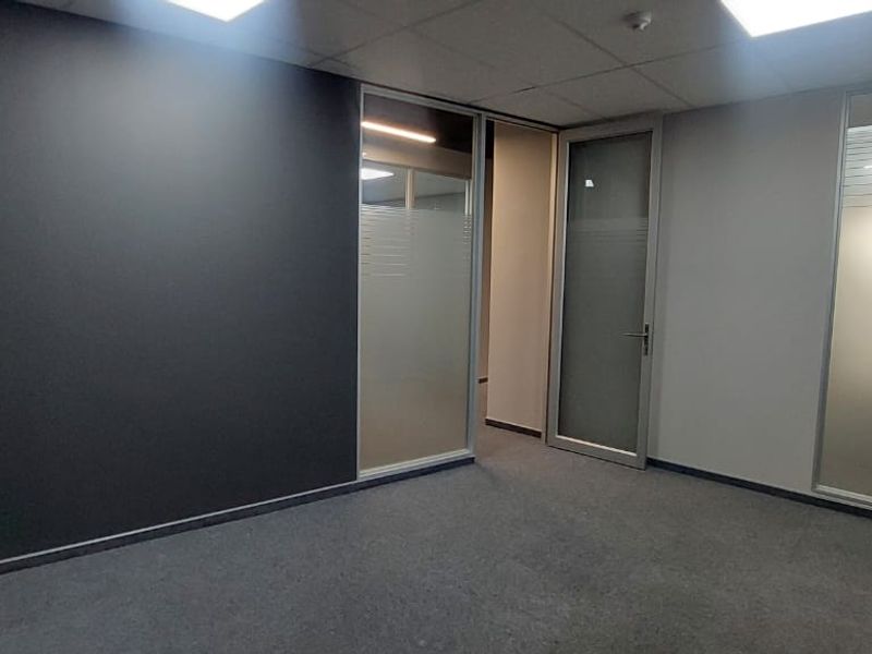 A modern, empty office space featuring glass partition walls, a frosted glass door, carpeted flooring, and a suspended ceiling with recessed lighting.