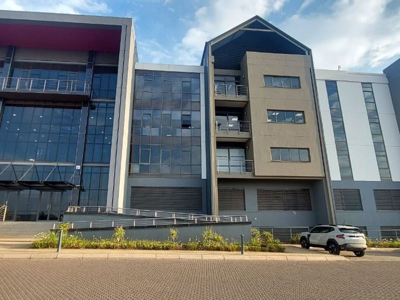Modern commercial office building featuring a striking glass facade, metal cladding, and integrated balconies with steel railings, set within a well-maintained landscaped environment.