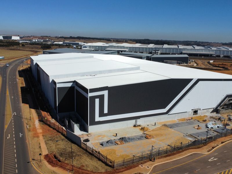 Aerial view of a large industrial warehouse facility featuring modern metal cladding, a pitched roof, and surrounding perimeter fencing in a developing industrial park.