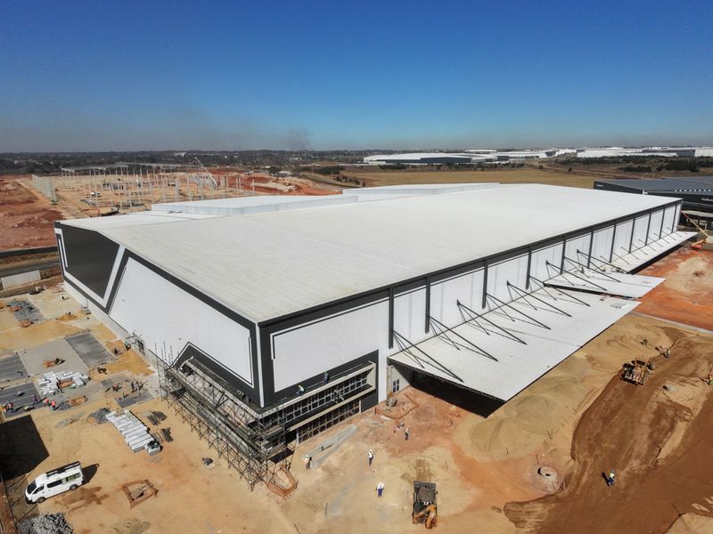Aerial view of a large-scale industrial warehouse facility under construction, featuring a expansive metal roof, cantilevered canopy, and surrounding site development.