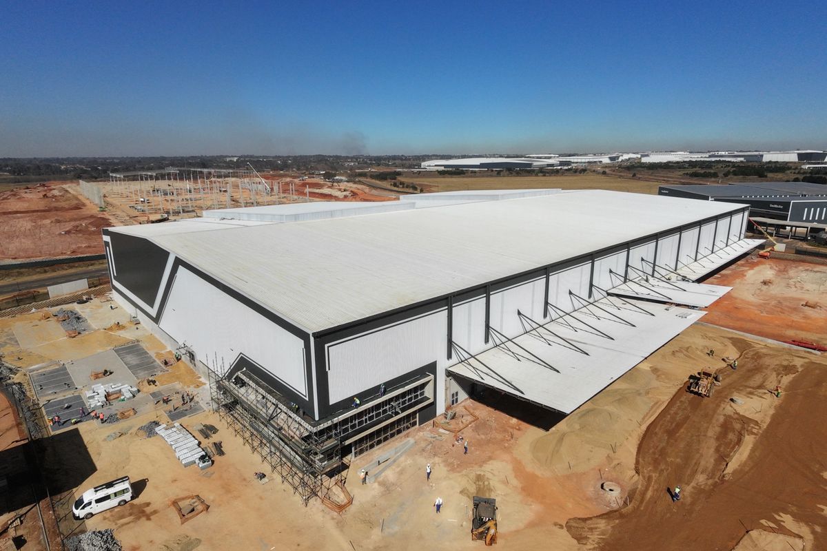 Aerial view of a large-scale industrial warehouse facility under construction, featuring a expansive metal roof, cantilevered canopy, and surrounding site development.