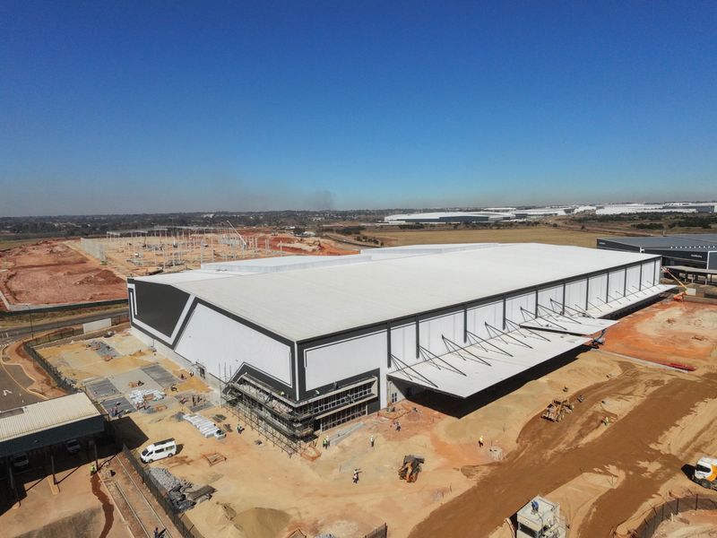 Aerial view of a large-scale industrial warehouse facility under construction, featuring extensive metal roofing, loading canopies, and surrounding site development.
