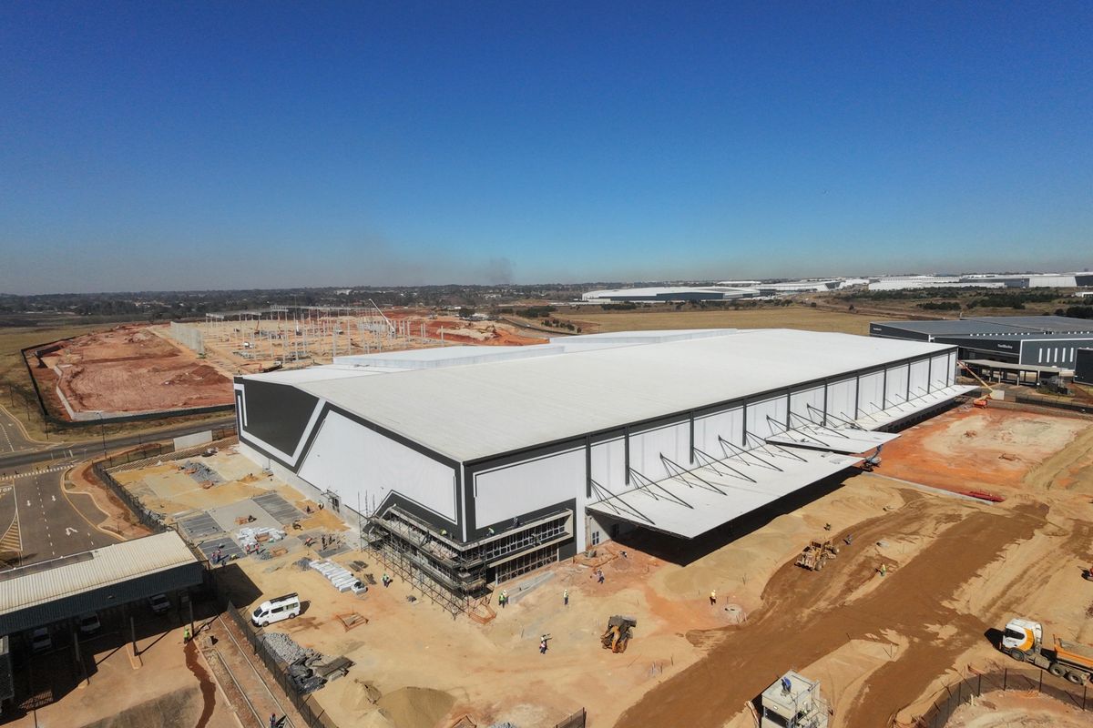 Aerial view of a large-scale industrial warehouse facility under construction, featuring extensive metal roofing, loading canopies, and surrounding site development.