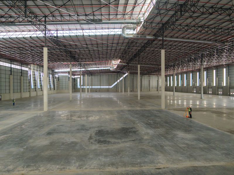 A large, open-plan industrial warehouse space featuring high ceilings with exposed steel trusses, concrete flooring, and integrated fire sprinkler and ventilation systems.