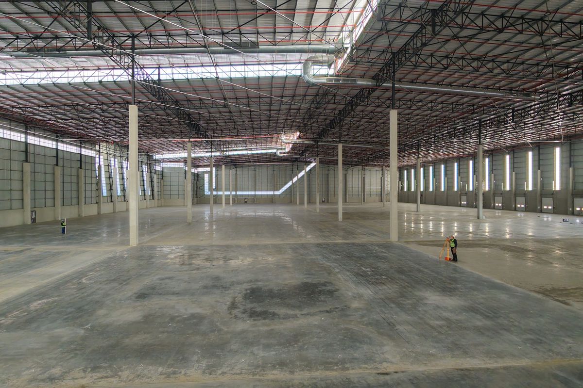 A large, open-plan industrial warehouse space featuring high ceilings with exposed steel trusses, concrete flooring, and integrated fire sprinkler and ventilation systems.