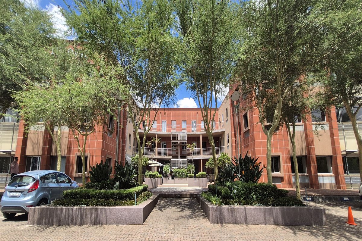 A well-maintained office building complex featuring a red brick facade, central courtyard access, and landscaped garden beds with paved driveway.